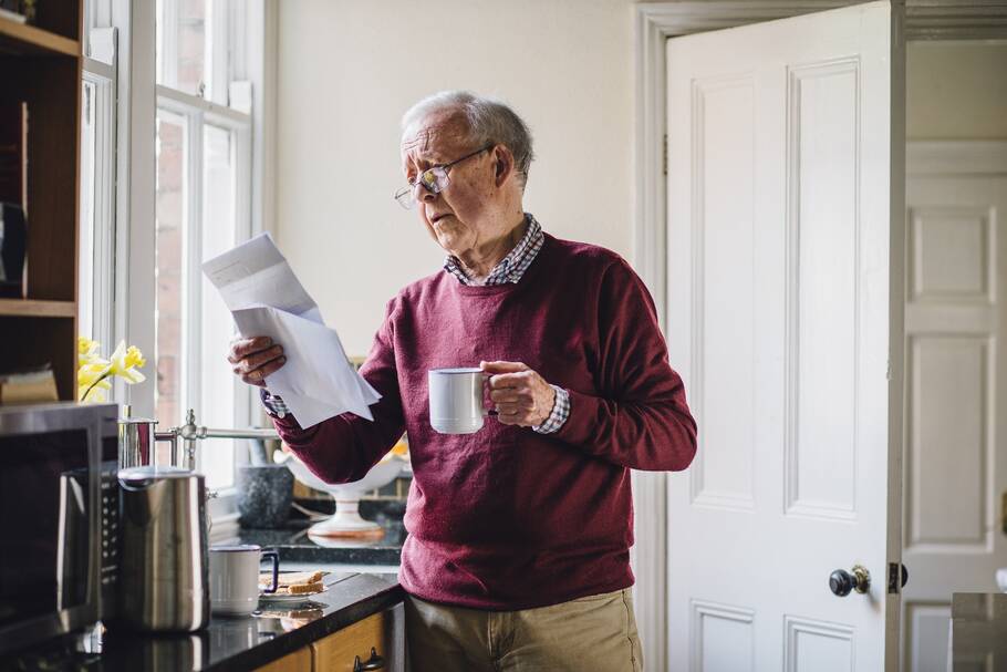 An elderly man stands by his kitchen counter, holding his cup while reading a document.